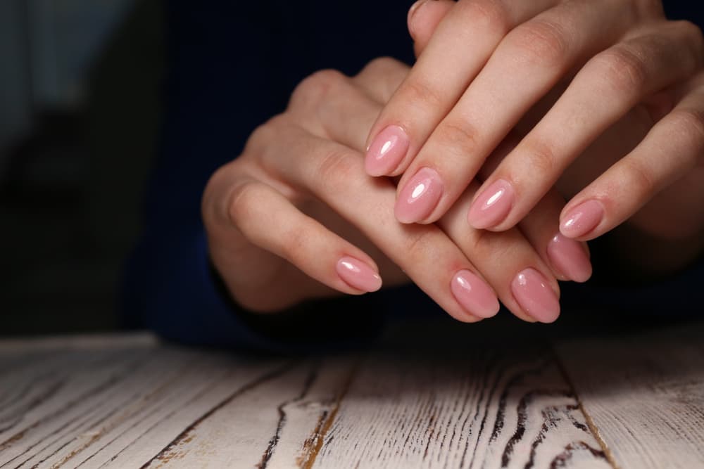 Close-up of hands with glossy pink BIAB nails resting on a wooden surface, featuring a soft oval shape and a smooth, high-shine finish.