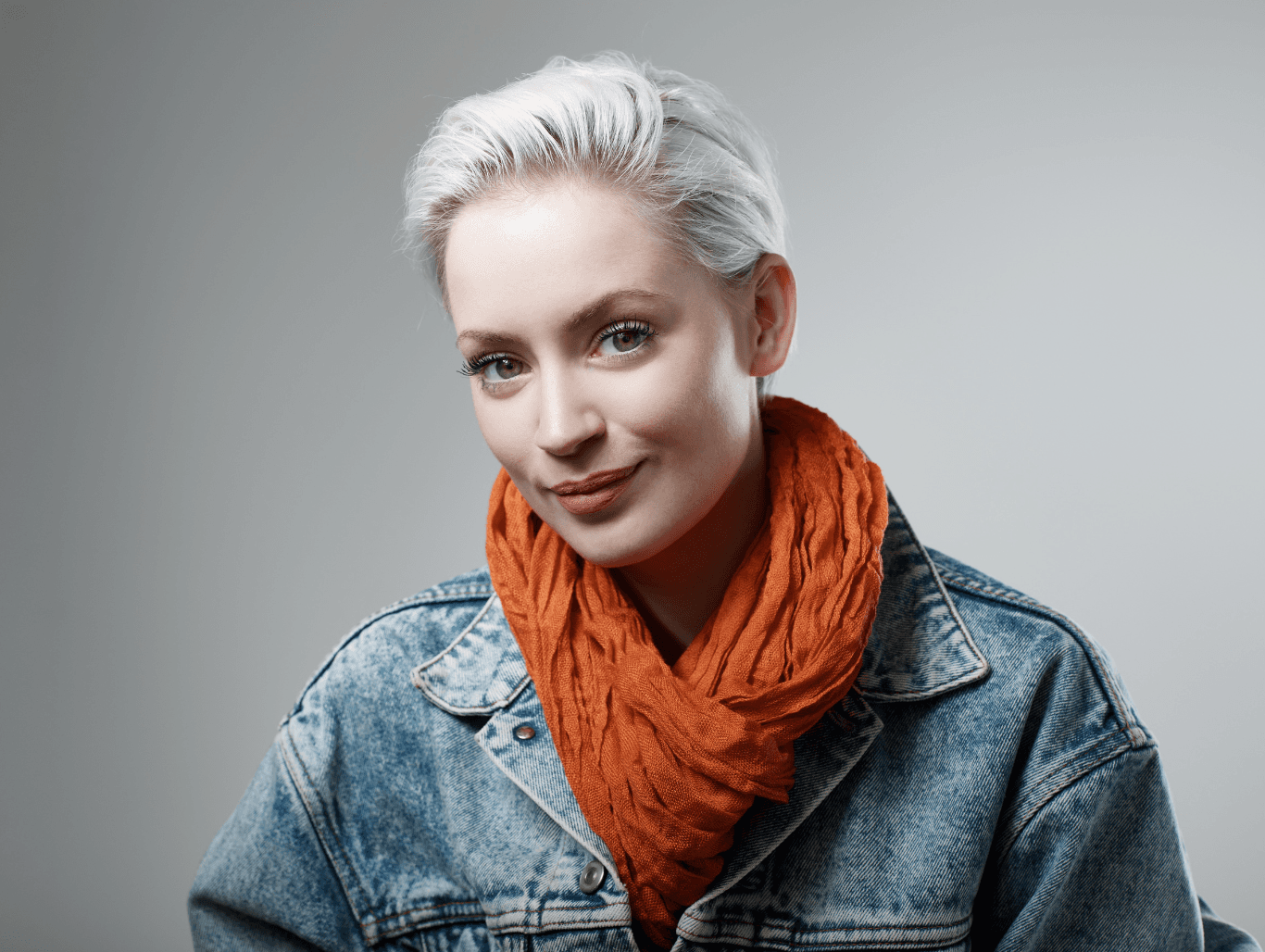 Woman with short platinum blonde hair and visible root regrowth, styled in a soft wave, wearing a denim jacket and orange scarf – representing 9+ weeks of bleach regrowth.